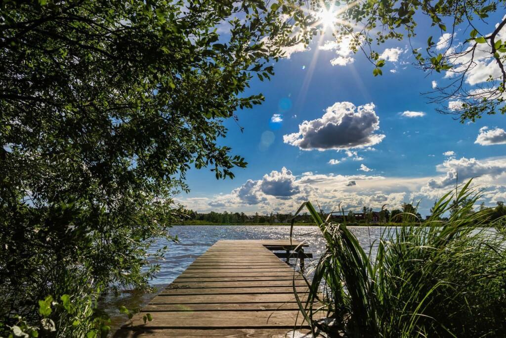 Kuvassa on aurinko, laituri, poutapilviä ja järvi.